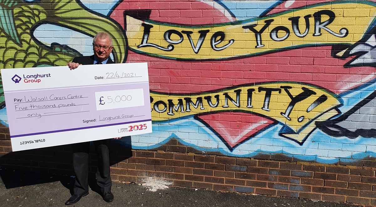 A man holding a cheque for Walsall Carers Centre, stood in front of graffiti that reads "love your community"