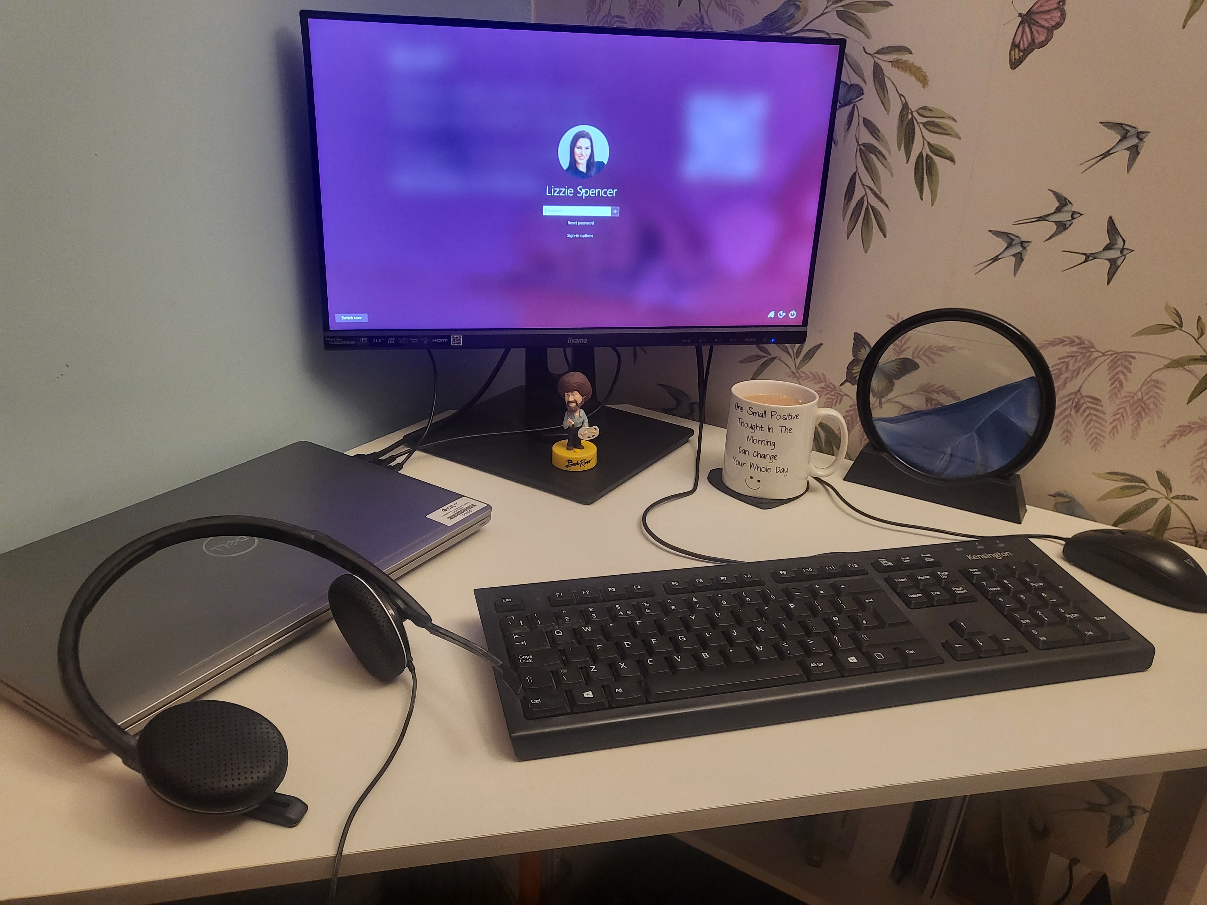 A work desk is neatly lined with a headset, keyboard, laptop, mouse, magnifying glass and a warm hot beverage. On the monitor stands a cute lego figure..